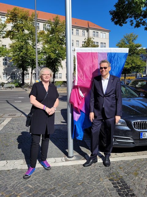 Bildvergrößerung: Eine Frau und ein Mann stehen vor einem Fahnenmast, an dem eine pink-lila-blaue Flagge hängt.