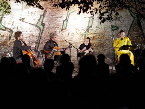 Bildvergrößerung: Draußen auf einem Platz in der Dunkelheit eine beluchtete Bühne mit drei Instrumentalist_innen in dunkler Kleidung und einer Sopranisten in einem goldenen Kleid. Im vordergrund das Publikum.