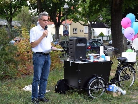 Bildvergrößerung: Ein Mann steht mit einem Zettel und einem Mikro auf einer Wiese und redet. Neben ihn steht ein Lastenfahrrad mit Luftballons.