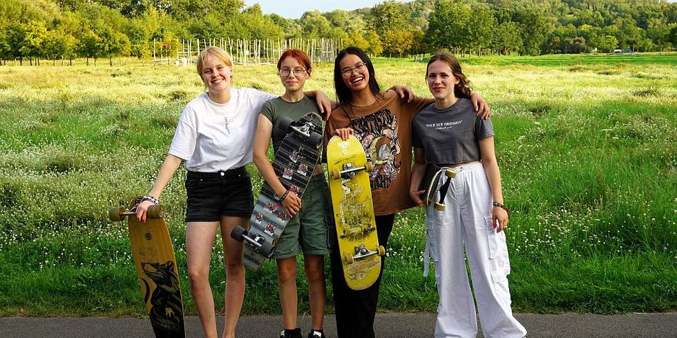 Das Pressefoto PULS-Camp der FreiwilligenAgentur Marzahn-.Hellersdorf zeigt vier lachende junge Frauen vor einer Grünfläche mit je einem Longboard in den Händen.