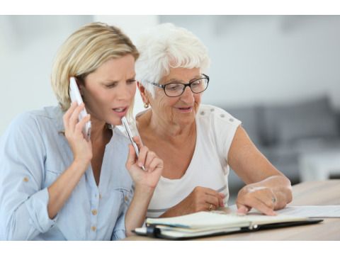Seniorin zeigt auf eine Seite in einem Terminkalender, eine junge Frau sitzt daneben und telefoniert