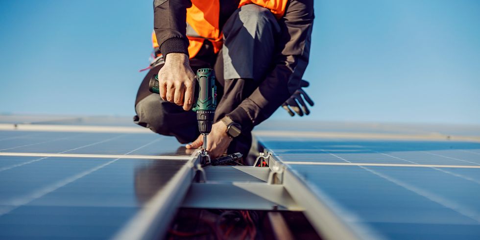 Handwerker mit Akkuschrauber auf Dach zwischen Solarmodulen