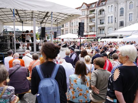 Bildvergrößerung: Viele Menschen stehen vor einer Bühne, auf der einige Personen Musik machen.