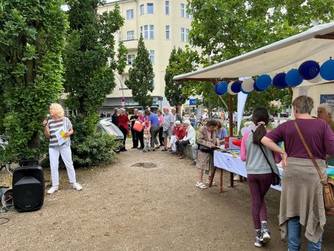 Bildvergrößerung: Eine Frau mit Mikrofon spricht zu Menschen auf einem Platz mit Marktständen. 