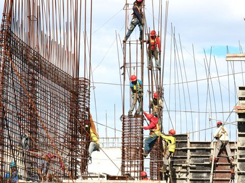 Arbeiter*innen bauen ein Gerüst auf in großer Höhe auf einer Baustelle im Freien.