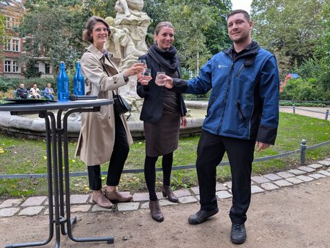 Bildvergrößerung: Zwei Frauen und ein Mann stehen vor einem Brunnen und halten jeweils ein Glas Wasser in der Hand.