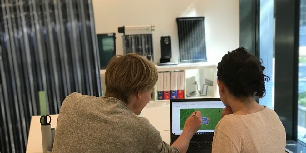 2 Personen sitzen am Schreibtisch. 1 Person berät dabei die andere Person. Beide sehen dabei auf den Laptop, der vor Ihnen steht.