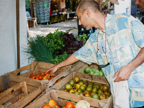 Ein Mann wählt Obst und Gemüse aus einer Holzkiste auf dem Wochenmarkt. Die Szene betont regionale und unverpackte Einkaufsmöglichkeiten als Beitrag zu Zero Waste.