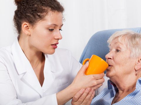 Junge Pflegerin führt einen Trinkbecher an die Lippen einer bettlägerigen Seniorin