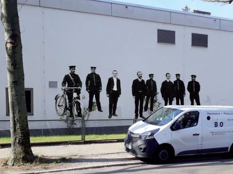 Bildvergrößerung: Auf der Fassade eines Gebäudes sind mehrere Männer abgebildet, einige davon in Post-Uniform, einer hat ein Fahrrad.