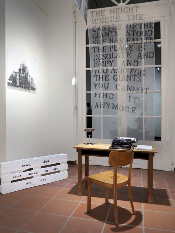 Ein Holztisch mit einer alten Schreibmaschine darauf steht vor einer Fenstertür, vor der ein transparenter Stoff mit einem darauf gedruckten Gedicht hängt