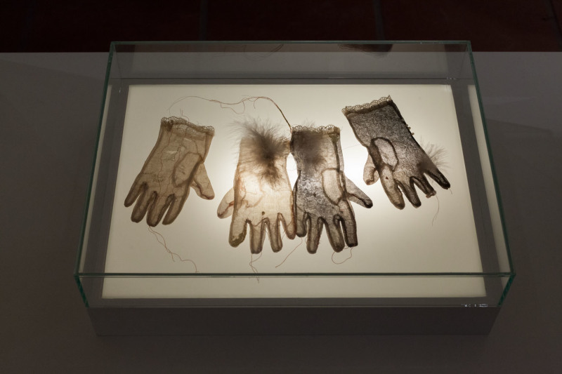 In einer Leuchtvitrine liegen zwei helle und zwei dunkle, dünne Handschuhe, durch die das Licht hinduchleuchtet. Diese bezeichnet die Künstlerin auch als Schmerzhandschuhe.