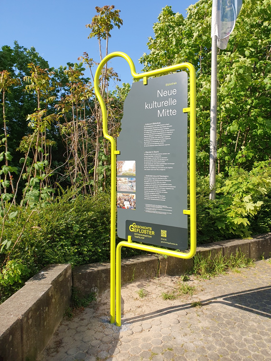 Stele Geschichtsgeflüster mit der Beschriftung Neue kulturelle Mitte auf einem Platz, im Hintergrund Bäume