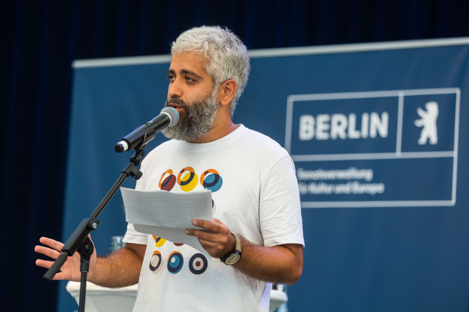 Mann mit Vollbart am Mikrofon mit Textblatt in der Hand 