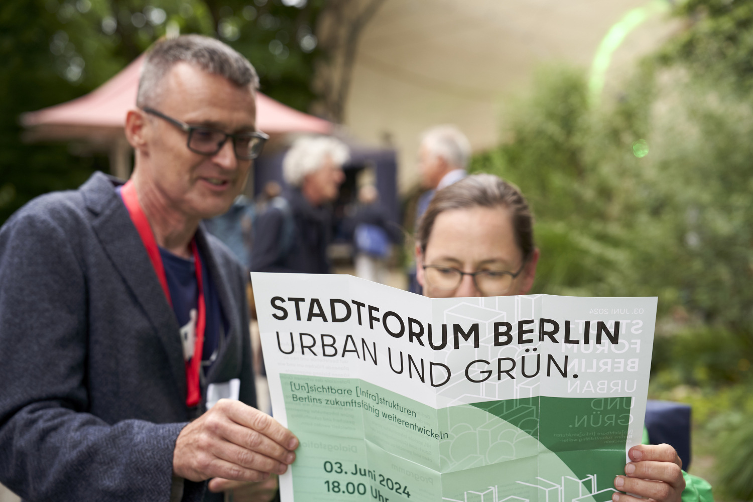 Ein Mann und eine Frau schauen sich ein Plakat an, dass die Frau vor sich in der Hand hält. Zu lesen ist „Stadtforum Berlin Urban und Grün.“