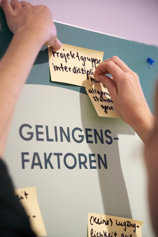 Eine Person pinnt zwei Post its an eine grüne Pinnwand auf der groß „Gelingensfaktoren“ steht. Auf dem Post it steht geschrieben „Projektgruppe interdisziplinär“