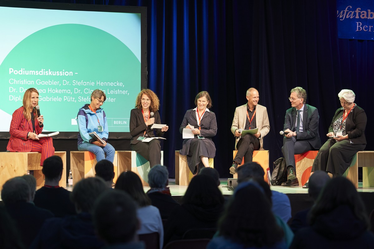 Sieben Personen sitzen auf einer Bühne. Im Hintergrund ist eine grüne Leinwand auf der „Podiumsdiskussion“ zu lesen ist. Auf dem Podium sitzen Dr. Dorothea Hokema, Bezirksstadträtin Dr. Claudia Leistner, Prof. Dr. Stefanie Hennecke, Stefan Richter, Senator Christian Gaebler und Dipl.-Ing. Gabriele Pütz.