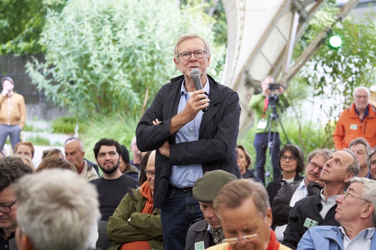 Ein Mann steht inmitten eines Publikums und spricht in ein Mikrofon, dass er in der Hand hält.