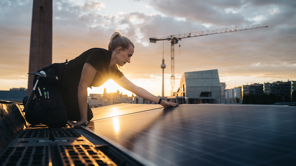 Handwerkerin bei Sonnenaufgang auf dem Dach, im Vordergrund Solarpanel, welches sie montiert. Im Hintergrund die Stadt Berlin.