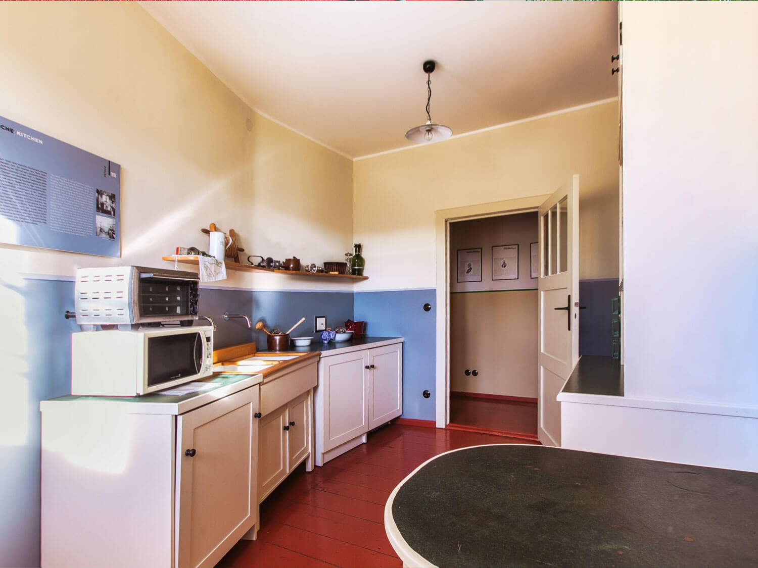 Rechts ein abgerundeter Tisch mit einer schwarzen Tischplatte. Links mehrere weiße Schränke. Auf dem vordersten Schrank steht eine Mikrowelle und ein kleiner siblerfarbener Backofen. Auf dem zweiten Schrank zwei eingelassene Spülbecken mit einer holzfarbenen Umrandung. Auf dem hintersten Schrank mehrere Schüssel und Töpfe. 